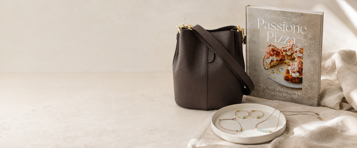 A dark brown leather bucket bag with gold-tone hardware sits beside a book titled 'Passione Pizza' and a small white dish with gold jewelry, arranged on a light beige surface with draped fabric.